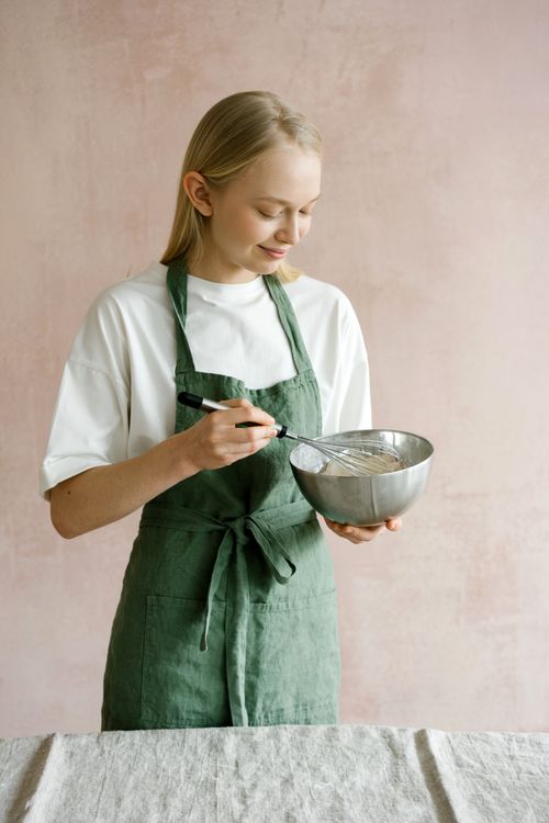 Free Caucasian woman in a green apron whisking ingredients in a metal bowl indoors. Stock Photo