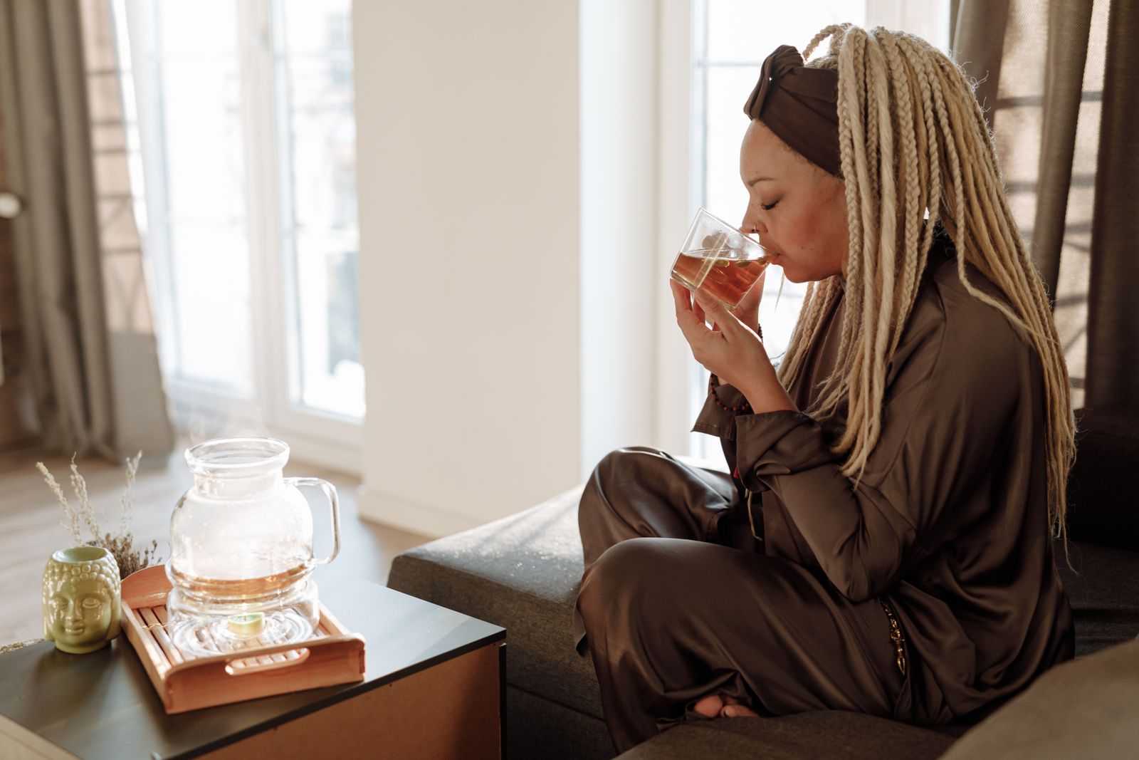 Woman with long blonde braids sipping tea from a glass cup on a sofa by a sunny window