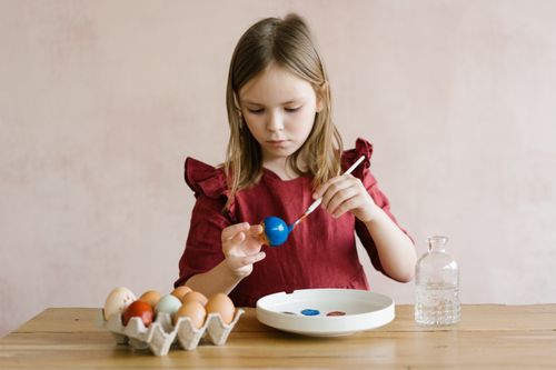 Free A young girl in a red dress painting Easter eggs at a wooden table, focused on creativity. Stock Photo
