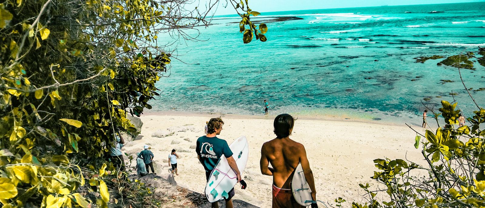 Surfers make their way down a lush path to a stunning beach in Bali, Indonesia.