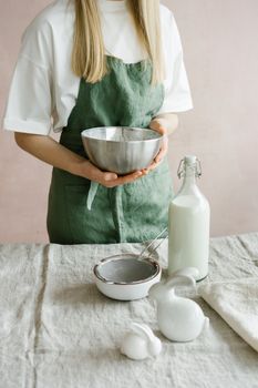 A woman in a green apron holds a mixing bowl with baking tools on a rustic table.