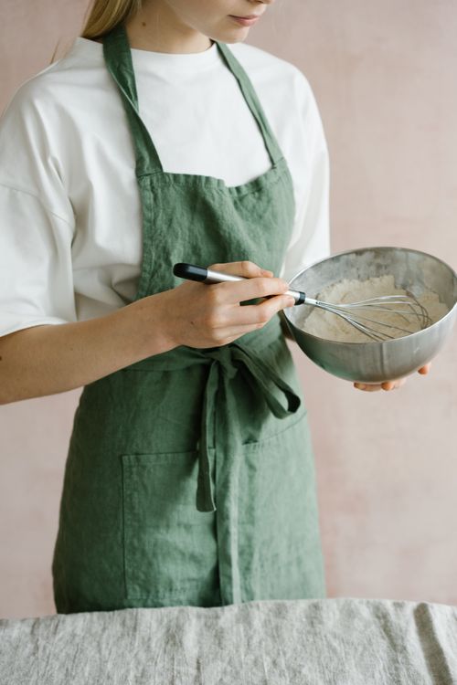 Free Woman in green apron whisking mixture in a steel bowl, illustrating home cooking. Stock Photo