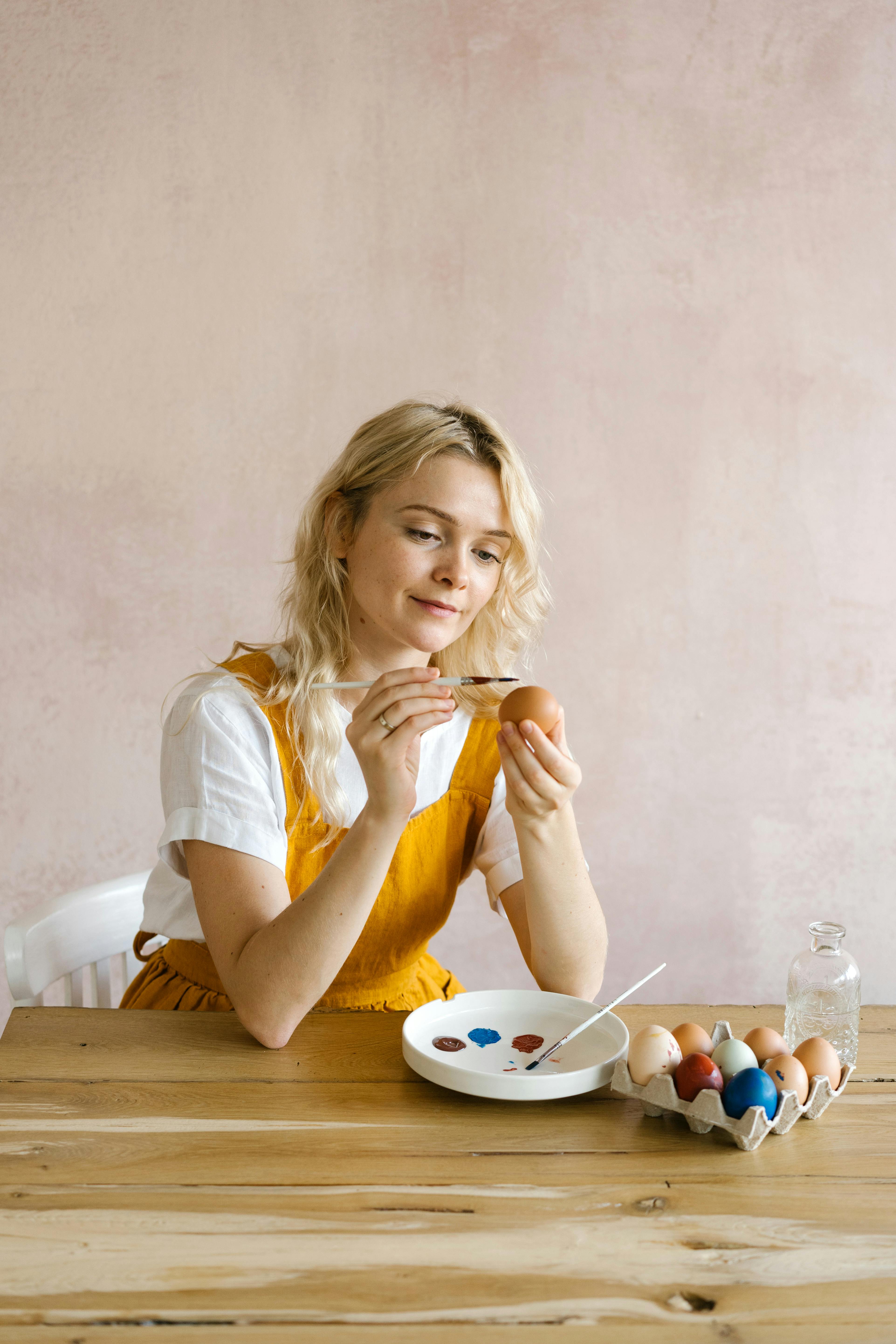 Woman painting Easter eggs with colorful paints on a wooden table indoors.