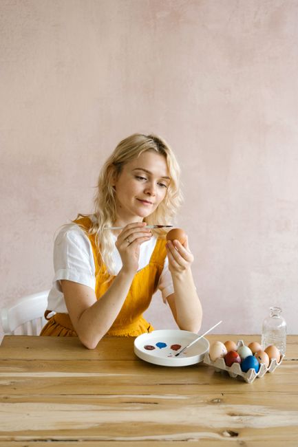 Woman painting Easter eggs with colorful paints on a wooden table indoors.