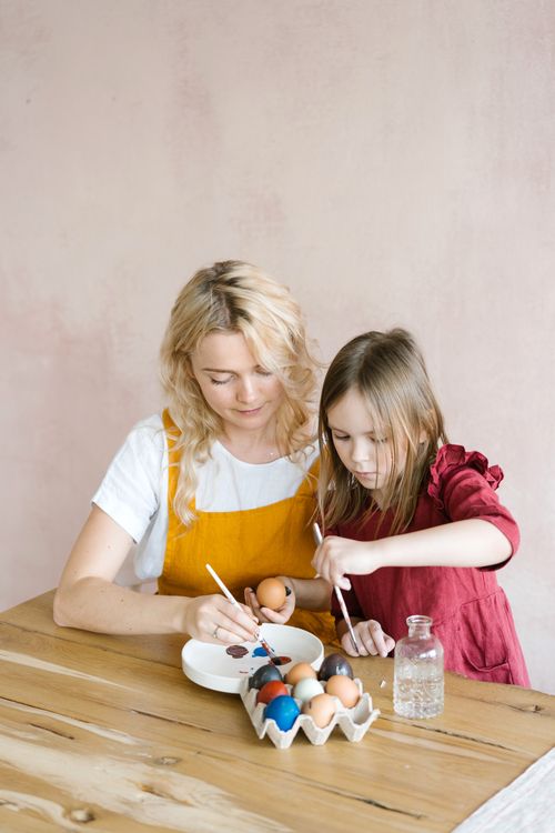 Free A mother and daughter joyfully painting Easter eggs indoors at a wooden table. Stock Photo