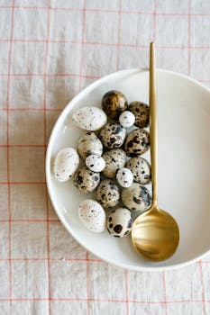 Minimalist top view of speckled quail eggs with a gold spoon on a white plate.
