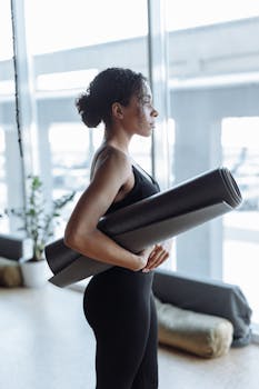 A woman in activewear holding a yoga mat in an indoor setting, ready for exercise.