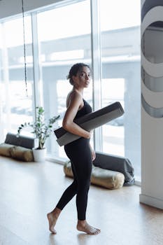 Black woman in activewear holding yoga mat indoors, embracing fitness and wellness.