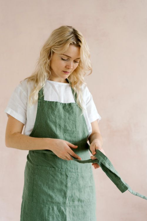 Free Blonde woman in a green apron adjusting it against a minimalist backdrop indoors. Stock Photo