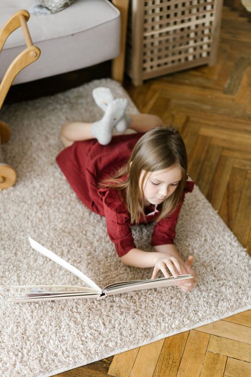 Free A young girl in a red dress lies comfortably on a rug reading a book in her cozy indoor space. Stock Photo