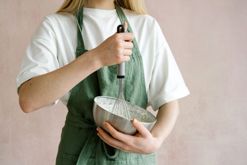 Close-up of a woman using a whisk in a stainless steel bowl, wearing a green apron.