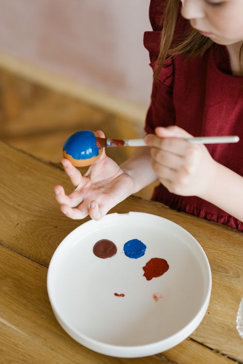 Free A young girl in a maroon dress painting a blue Easter egg with a brush on a wooden table. Stock Photo