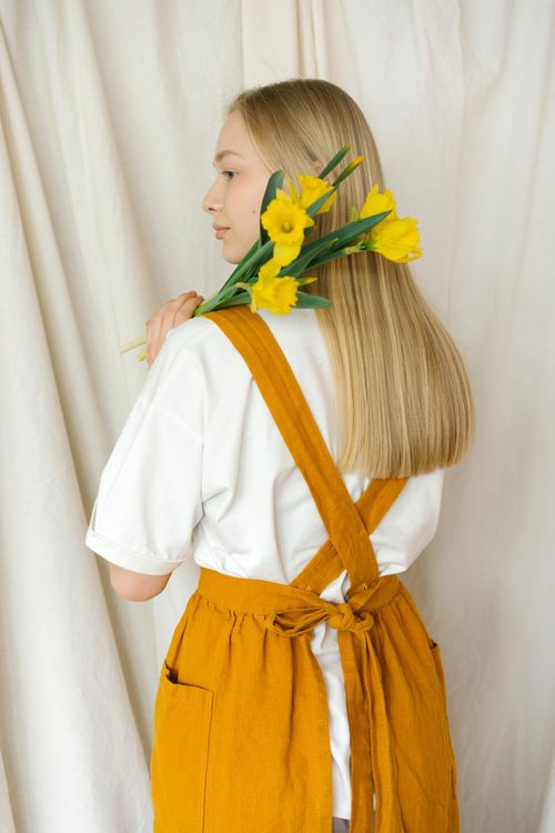 Free Back view of a woman in a mustard apron holding yellow daffodils against a beige curtain. Stock Photo