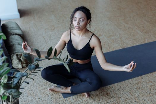 Young black woman sitting cross-legged in peaceful yoga meditation indoors. Relaxation and wellness concept.