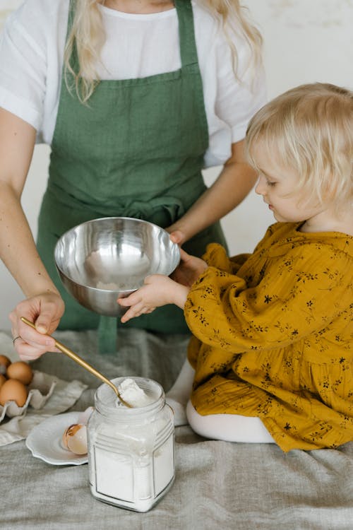 Free High angle of little daughter sitting on table with stainless bowl helping mother in making dough Stock Photo