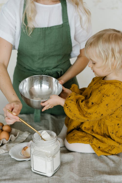 High angle of little daughter sitting on table with stainless bowl helping mother in making dough