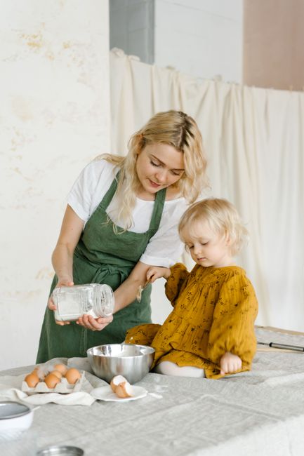 Focused mother in apron putting flour into metal bowl while adorable toddler watching on table in kitchen