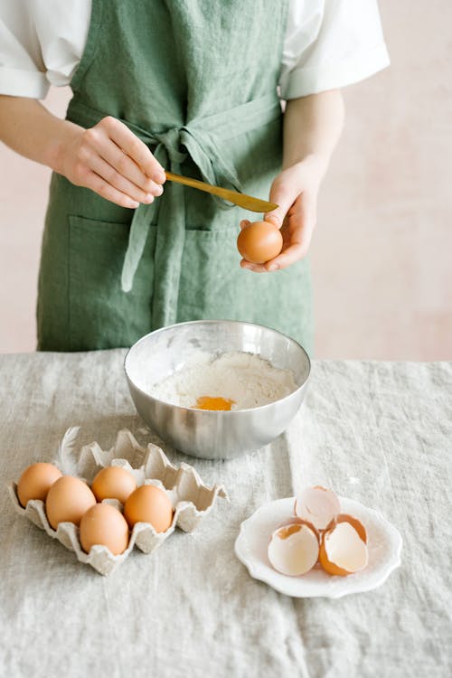 Crop unrecognizable woman in apron breaking fresh eggs while preparing food in light room in daylight