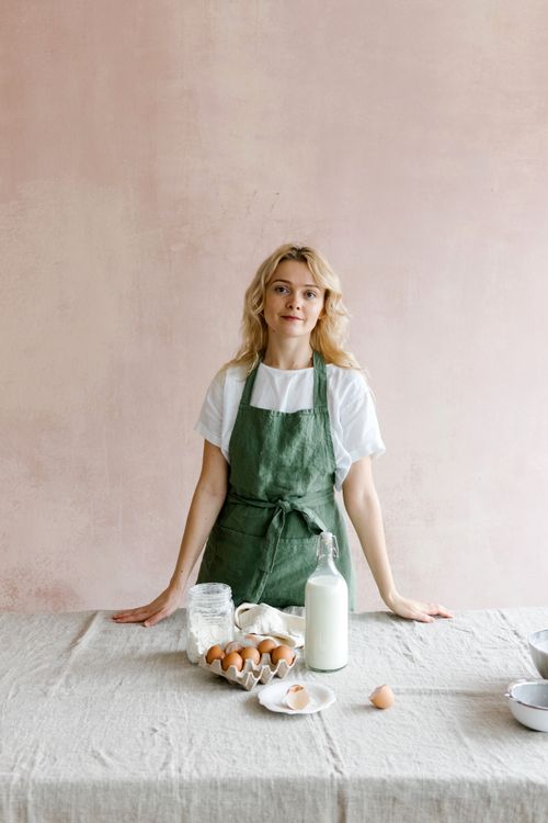 Free Charming female with blond hair in casual clothes and apron standing at table with eggs and bottle of milk in light room Stock Photo