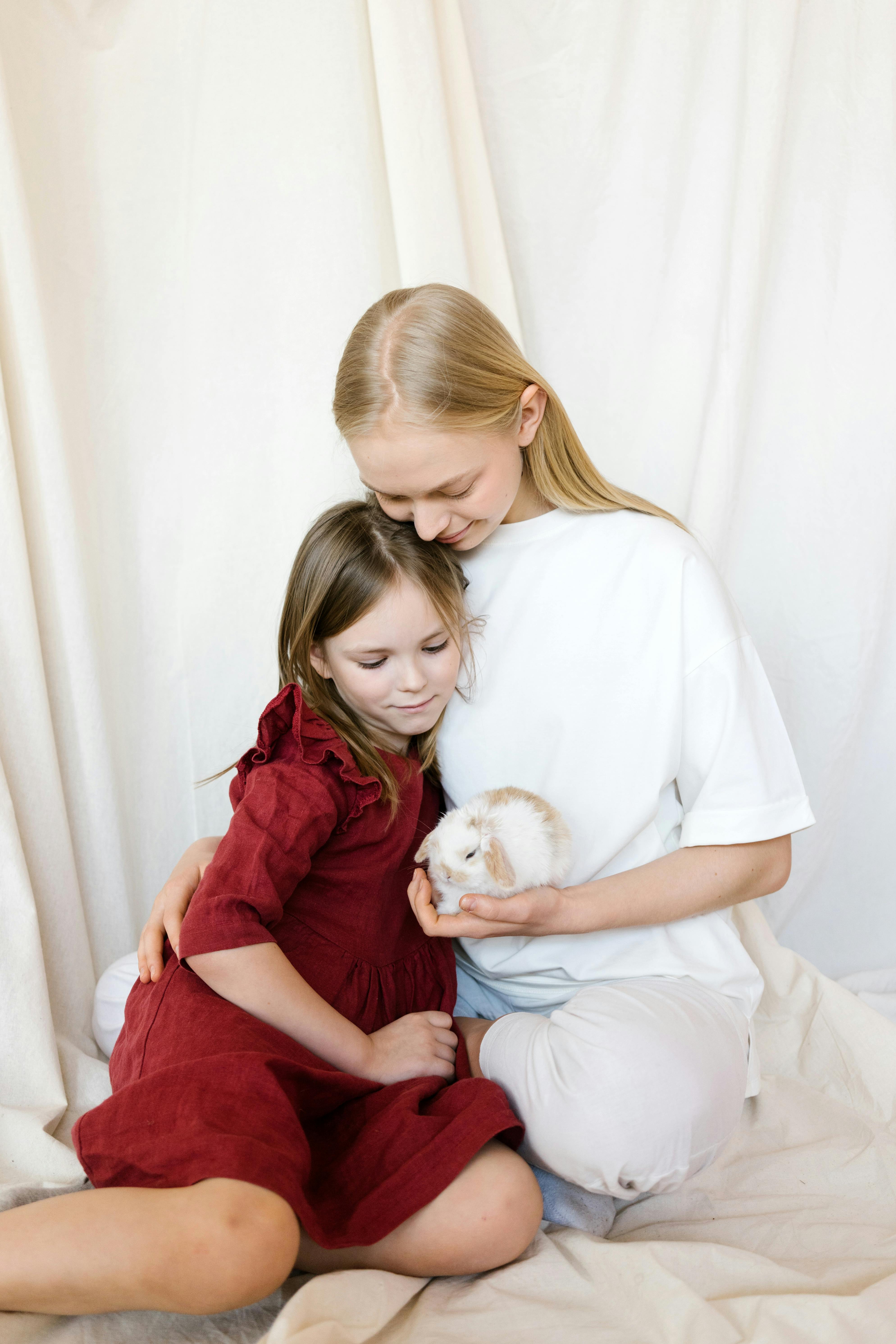 sisters embracing and looking at small rabbit against light curtain