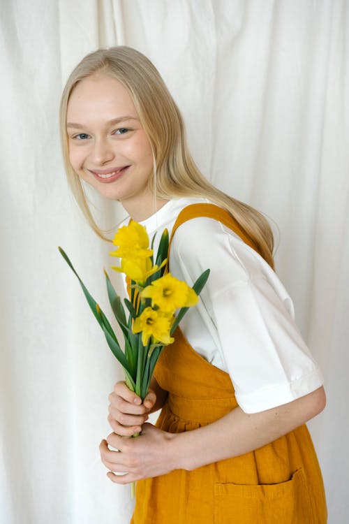 Cheerful young girl with blond hair in casual clothes standing with bouquet of yellow daffodils and looking at camera against light curtain