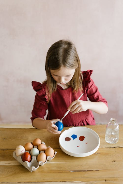 Free Calm girl with brown hair in casual clothes sitting at table and painting Easter eggs at home Stock Photo