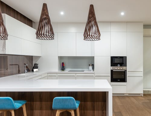 Turquoise chairs at counter with white cupboards and modern kitchenware in spacious light kitchen with hanging lamps in contemporary apartment