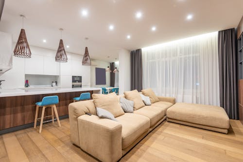 Comfortable sofa with cushions placed near kitchen counter with white cupboards and modern kitchenware in stylish light apartment with window