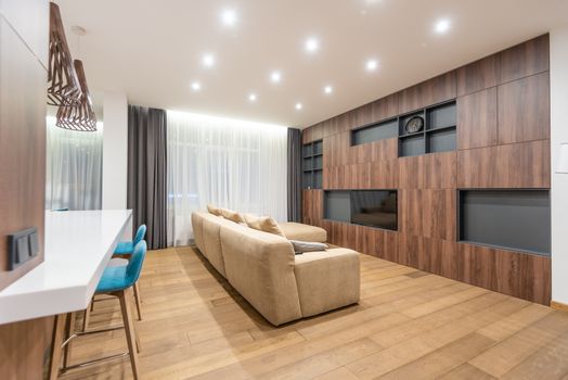 Chairs at white counter placed in spacious apartment with comfortable sofa and wooden cupboards with black shelves near big window