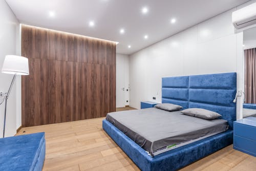 Comfortable blue bed with gray bedclothes placed between bedside tables in modern light spacious bedroom with wooden wardrobe at home