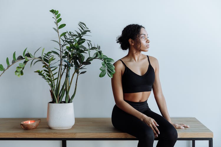 A Woman In Black Active Wear Sitting On A Wooden Table Beside An Indoor Plant And Lighting Candle