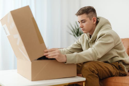 Free Man Sitting on Couch Opening a Cardboard Box on a Table Stock Photo