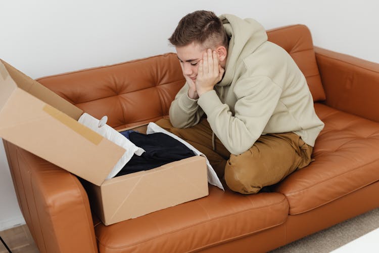 A Young Man In Sweater Sitting On A Couch While Looking At The Box With Clothes