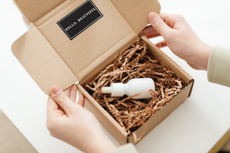 Hands Of A Person Opening A Box With A White Bottle