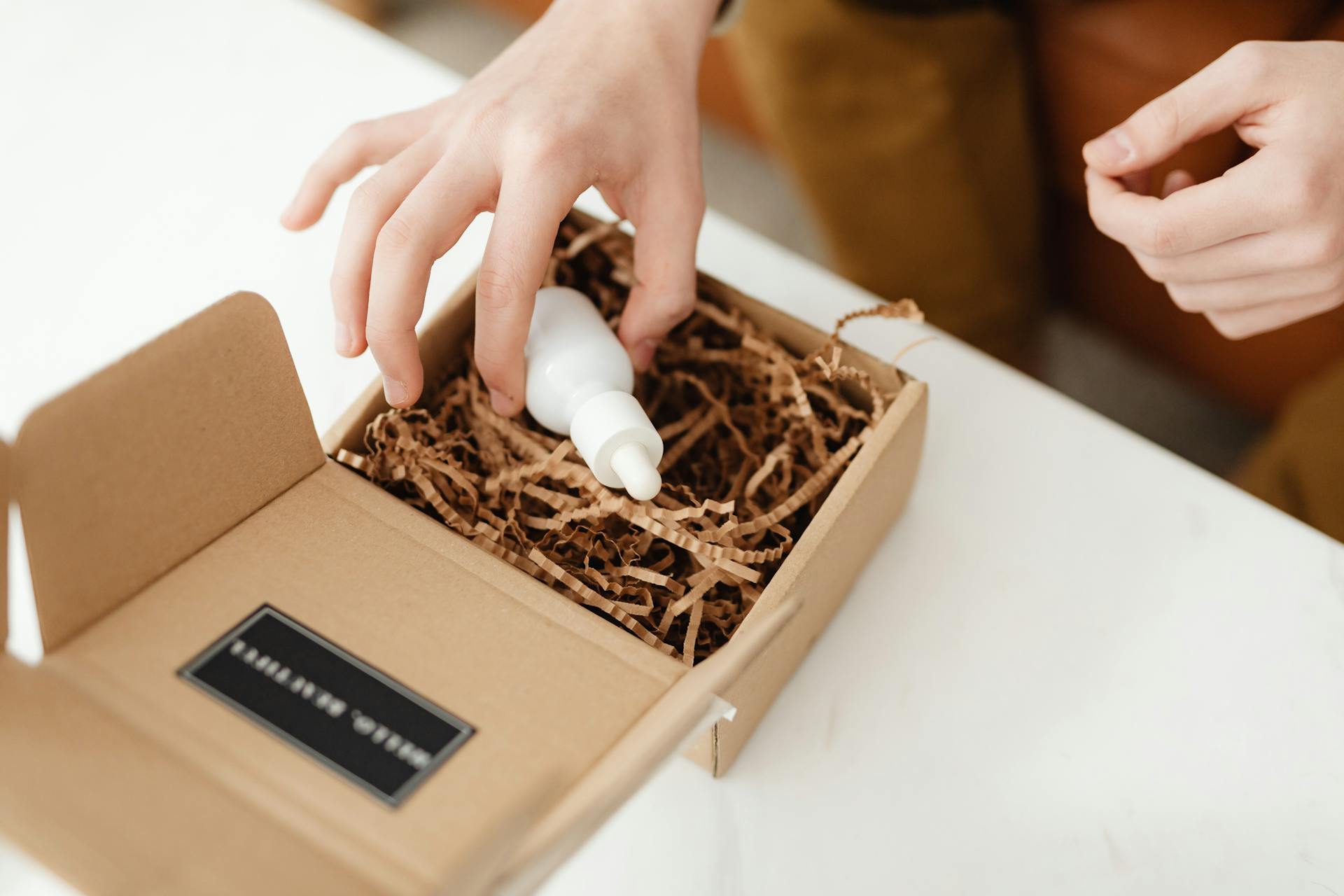 Hands unboxing a white dropper bottle from a cardboard package with shredded paper.