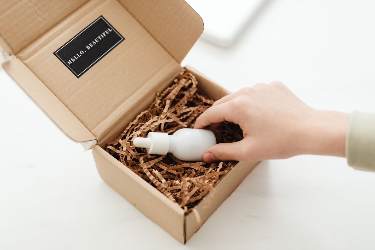 A hand placing a white dropper bottle into a kraft mailer box filled with paper crinkle fill