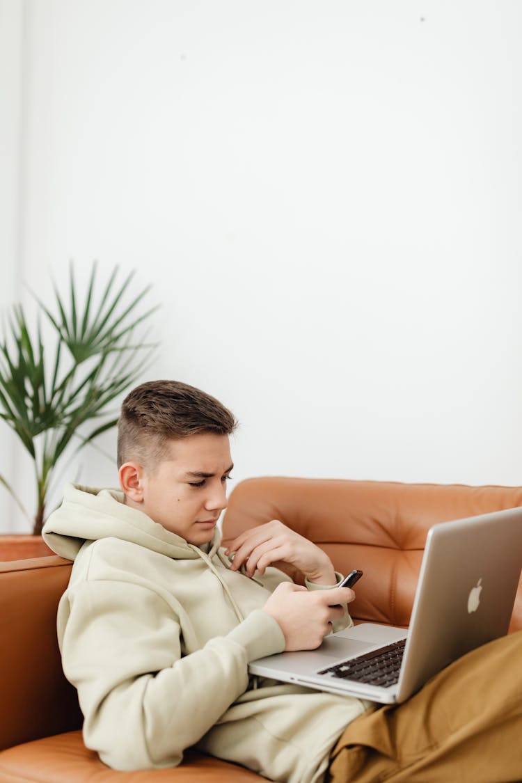 A Young Man In Hoodie Sweater Lying On The Couch While Looking At His Laptop