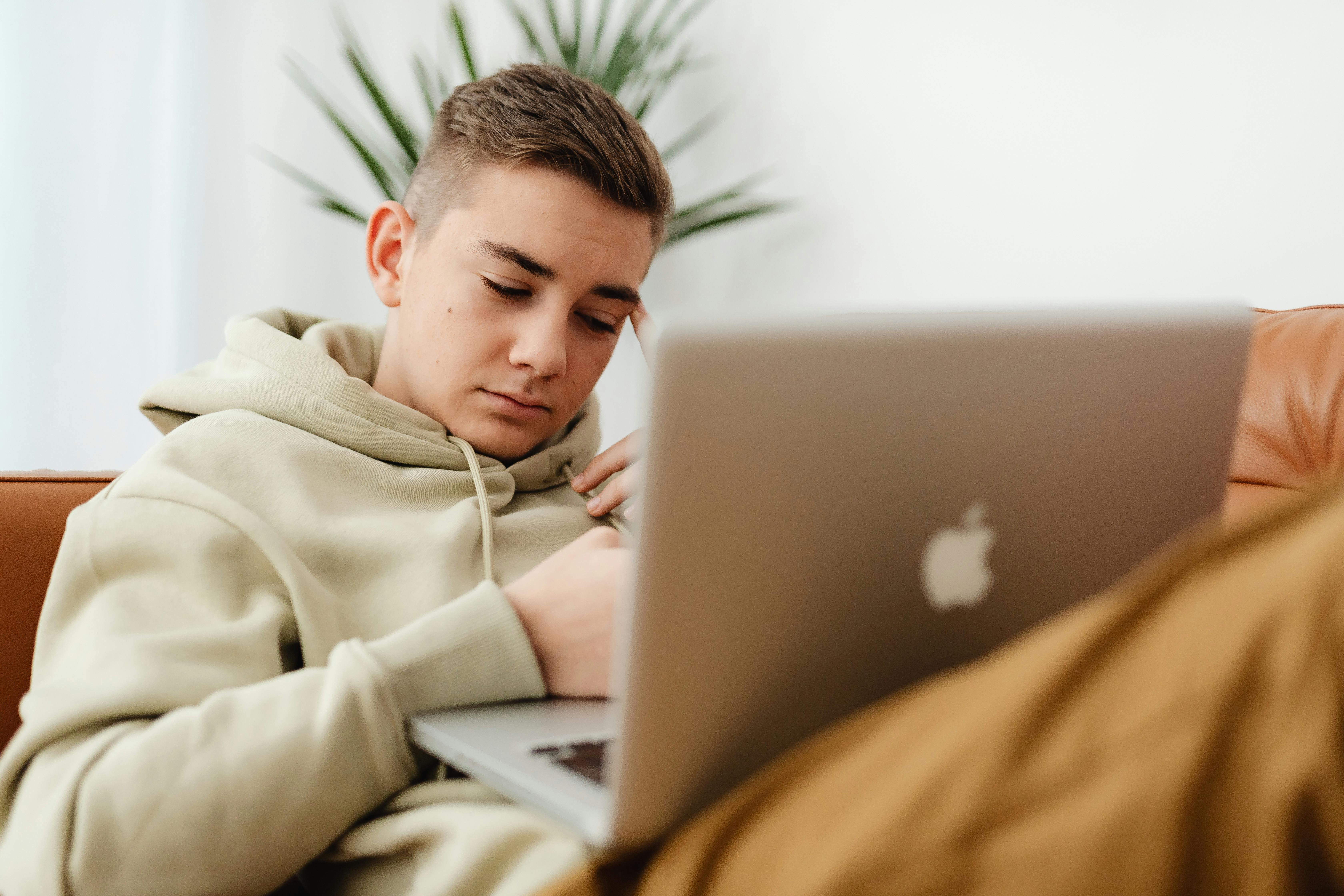 A young man in a hoodie relaxing on a sofa using a laptop with a focused expression.
