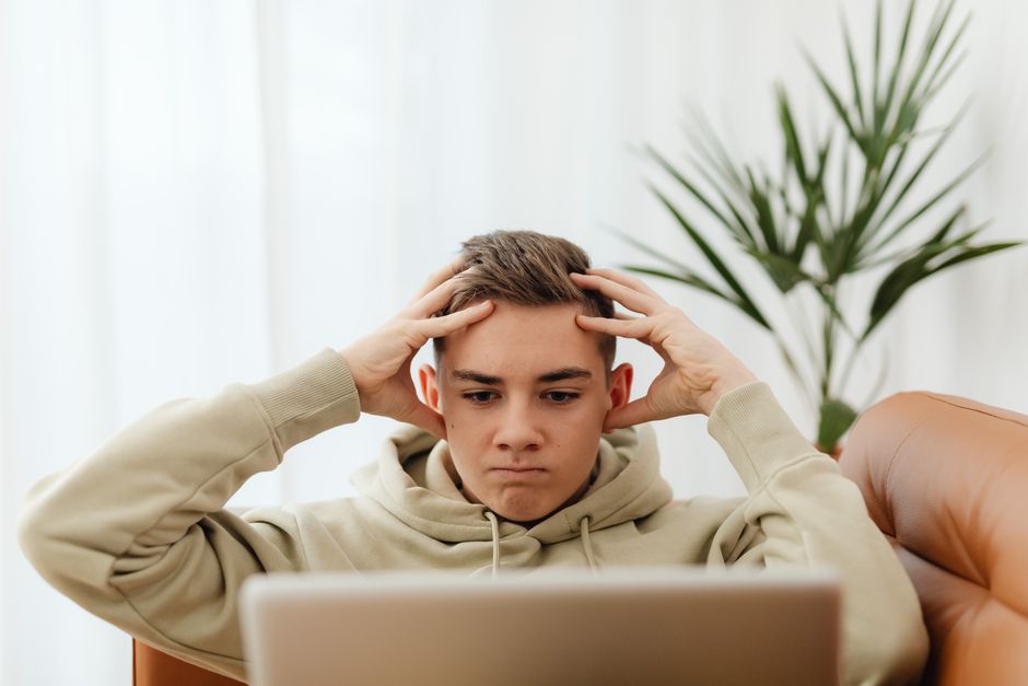A young man in a hoodie concentrates on his laptop, seated indoors, conveying stress or frustration.