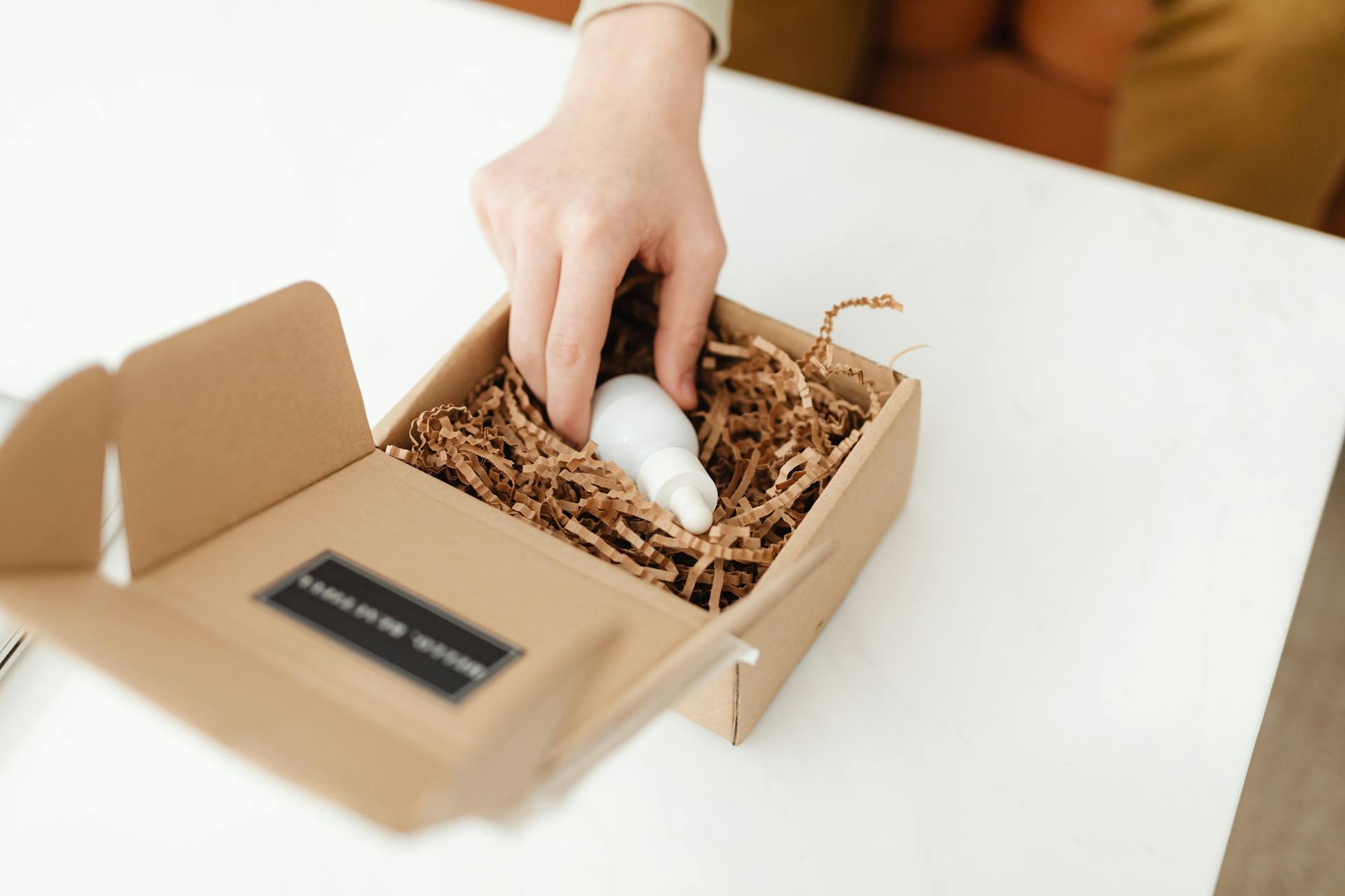 Close-up of a hand unpacking a cardboard box with a product bottle inside on a white table.