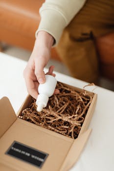 A person holding a dropper bottle over an open cardboard box with packing material inside.