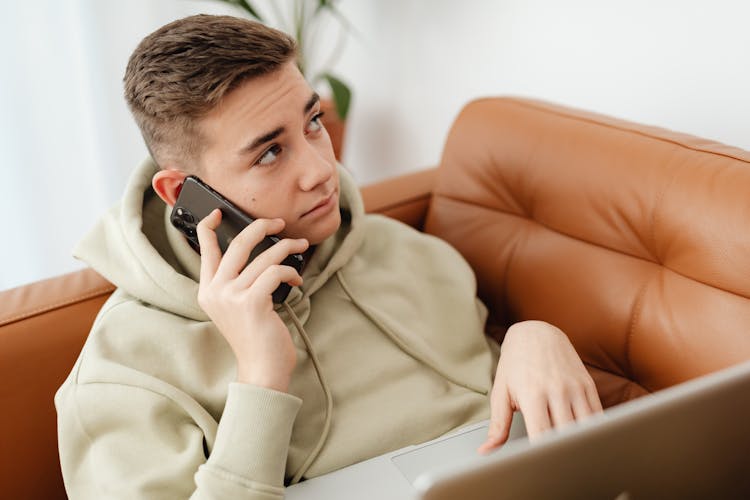 Man In Beige Hoodie Using Cellphone While Lying On Brown Sofa With Laptop 