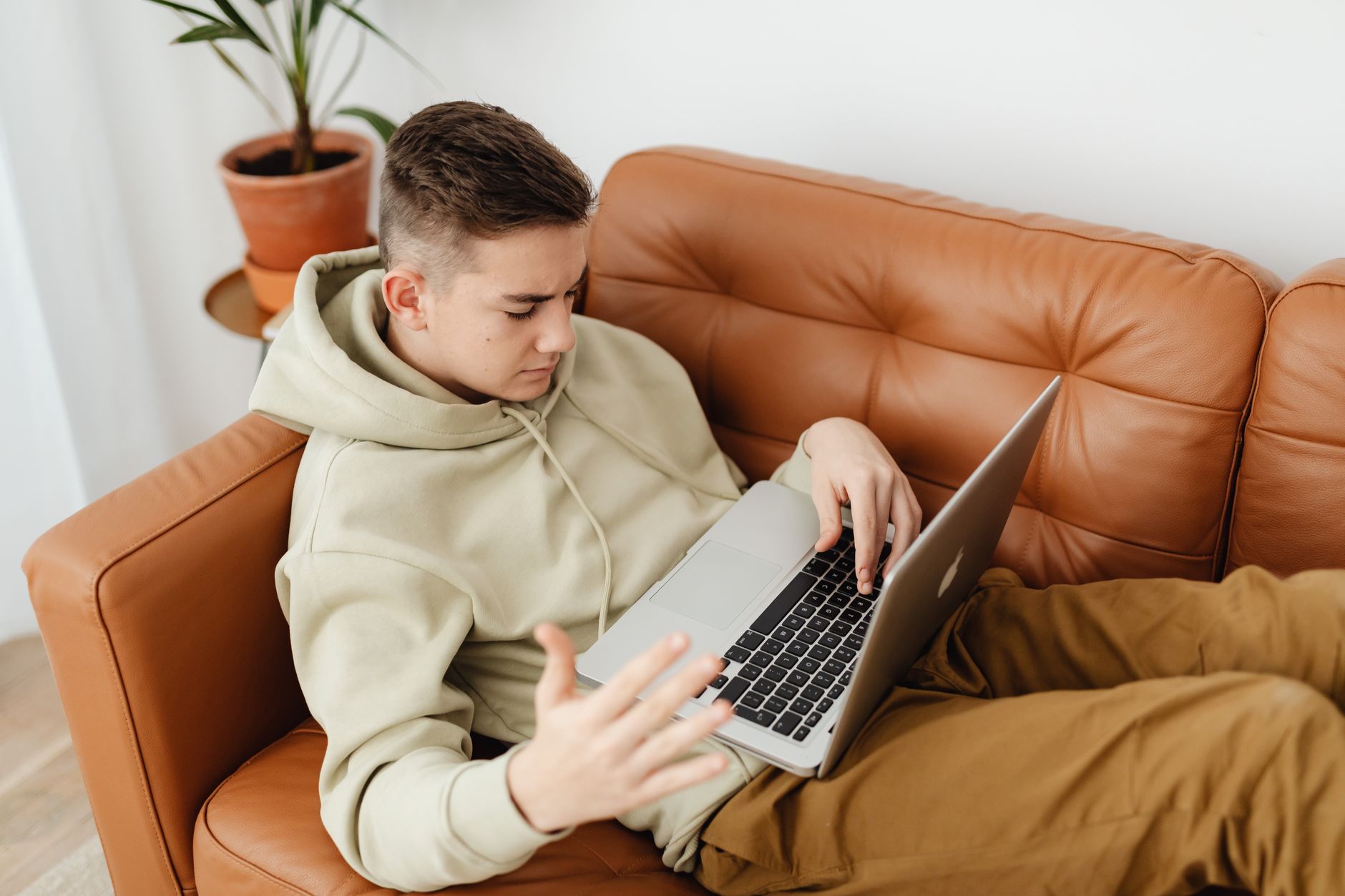 Junger Mann im Kapuzenpullover liegt auf einem Ledersofa, benutzt einen Laptop und gestikuliert