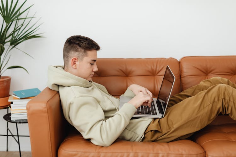 A Young Man Resting On A Couch While Typing On His Laptop