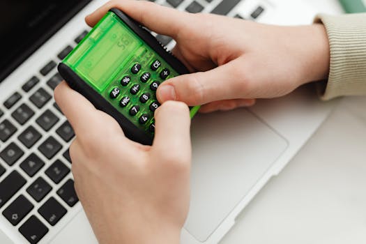 Close-up of hands using a green calculator on a laptop keyboard, indoor setting.