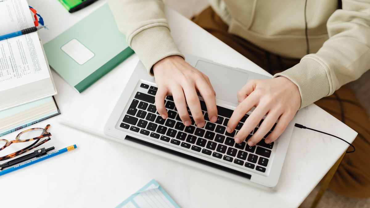 Close-up top view of hands typing on a laptop at a home desk surrounded by study materials.