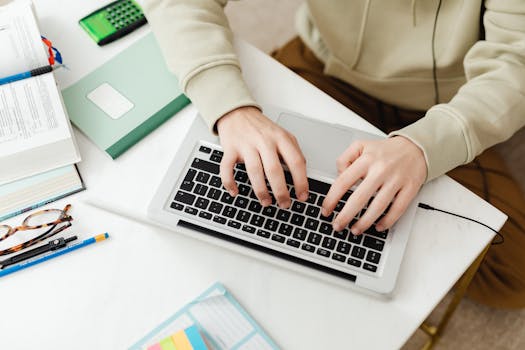 Close-up top view of hands typing on a laptop at a home desk surrounded by study materials.