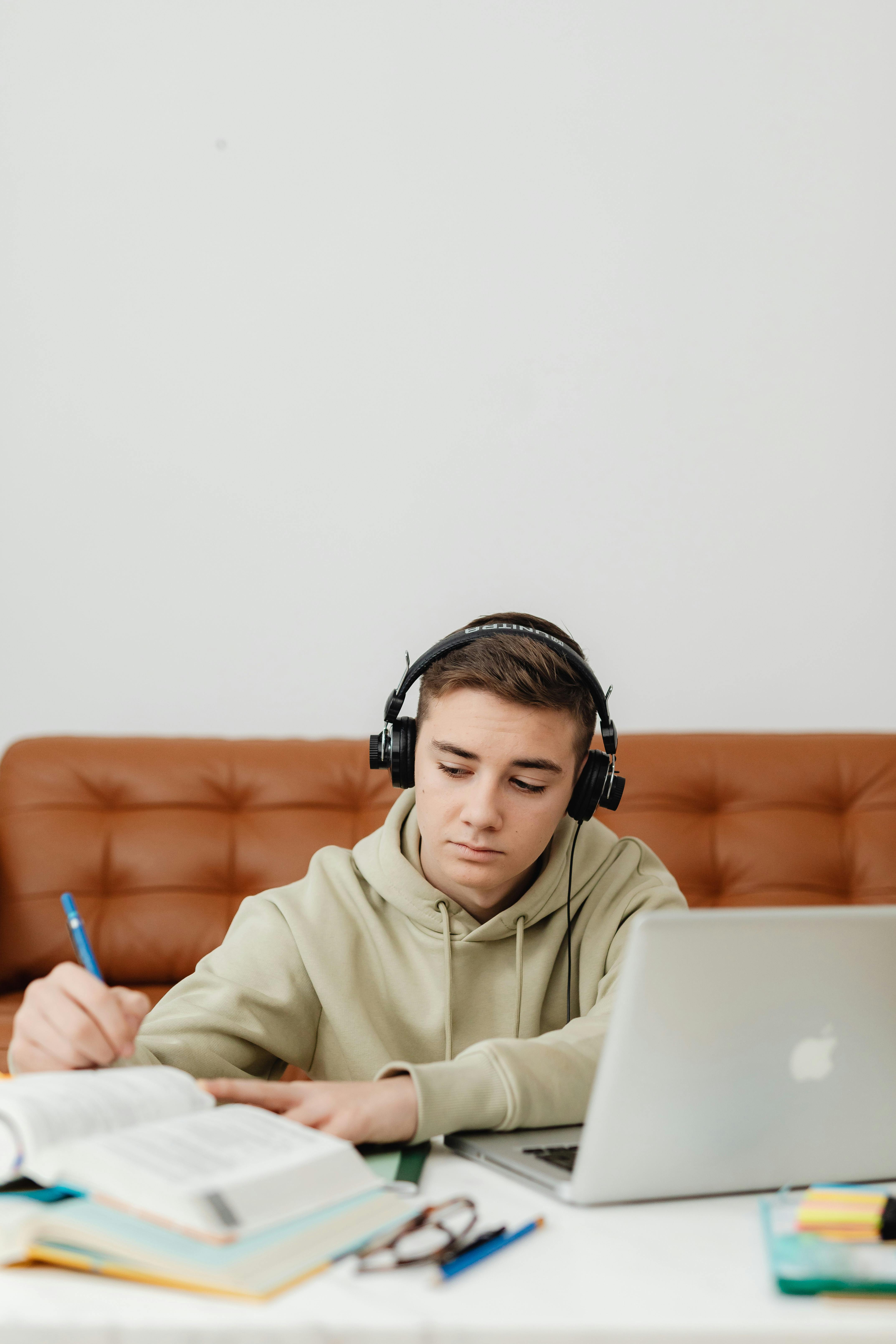 Teenage boy studying at home with headphones, laptop, and books. Focused on writing.