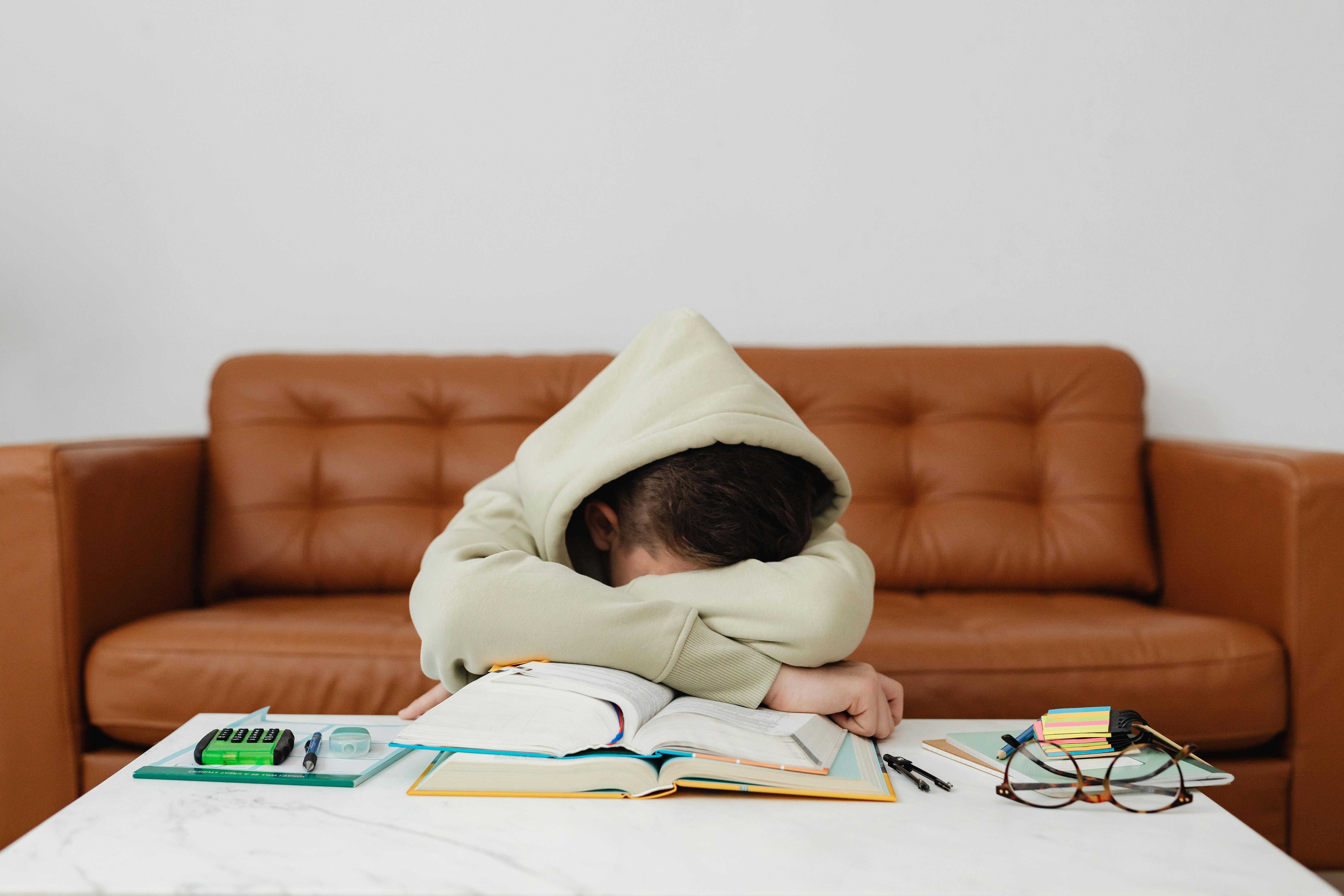 Free Child in Beige Hoodie Leaning Forward On Table Feeling Exhausted While Studying Stock Photo