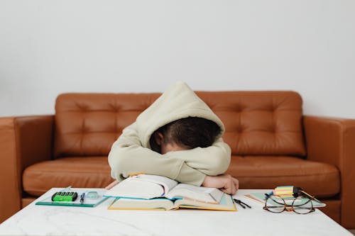 Child in Beige Hoodie Leaning Forward On Table Feeling Exhausted While Studying